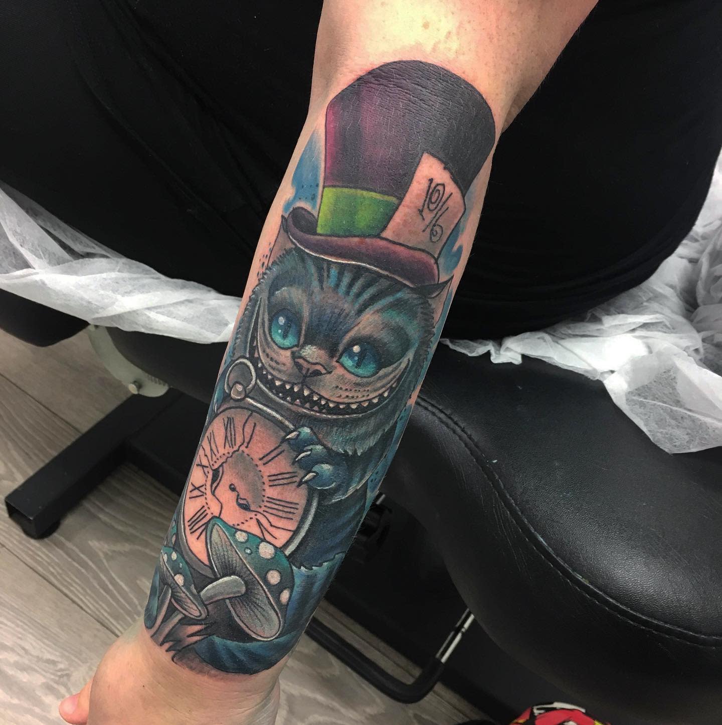 Colorful tattoo of the Cheshire Cat wearing a Mad Hatter hat, holding a cracked pocket watch with mushrooms at the base
